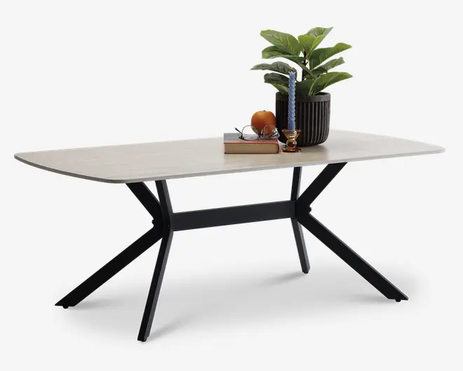 Rektangulært bord med en lys træbordplade og sorte metalben, der er vinklet udad for stabilitet. Bordpladen er dekoreret med en sort rillet urtepotte med en grøn plante, et blåt snoet stearinlys i en guldfarvet stage, en stak bøger, et par briller og en appelsin. Bordet har et moderne, minimalistisk design med rene linjer og en solid ramme.