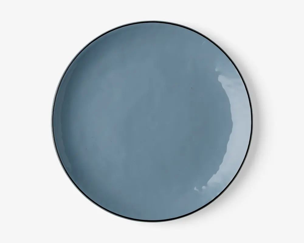 En rund, flad tallerken med en glat, blank overflade i en afdæmpet blå farve. Tallerkenen har en let hævet kant og et minimalistisk, moderne design. Overfladen er jævn og reflekterende med diskrete variationer i glasuren, som giver karakter.