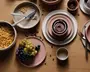 Et sæt keramikservice i afdæmpede rosa og beige nuancer, herunder tallerkener, skåle og kopper, arrangeret på et træbord. Sættet har et moderne, minimalistisk design med glatte, matte overflader. Nogle tallerkener er stablet, og skåle er sat ind i hinanden. Service vises med madvarer som druer, pasta og kaffebønner, hvilket fremhæver dets anvendelse til servering af forskellige retter.