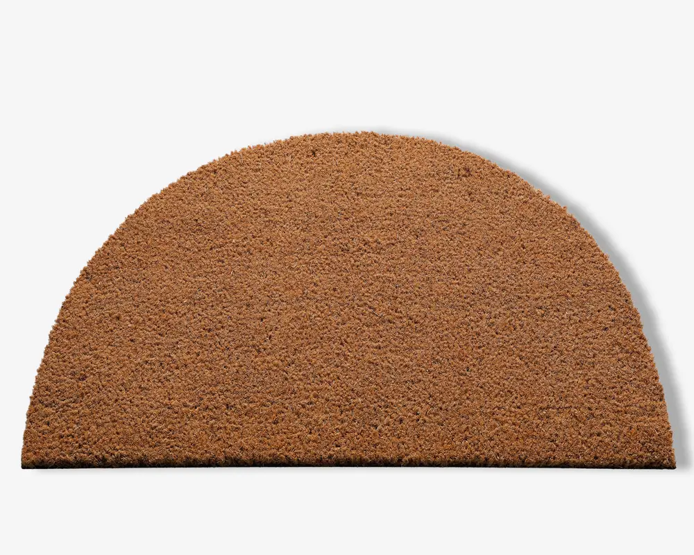 En halvcirkelformet måtte lavet af grove, naturlige brune fibre. Overfladen har en ru tekstur, der egner sig til at skrabe snavs af sko. Kanterne er pænt skåret, og måtten har en ensartet farve og tykkelse over hele fladen.