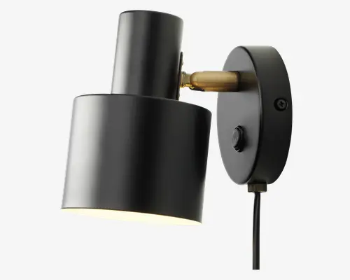Væghængt lampe med en mat sort cylindrisk metalskærm og base. Lampen har en messingfarvet justerbar arm, så lysets retning kan ændres. Der er en rund tænd/sluk-knap på basen, og en synlig sort ledning går ud fra bunden. Skærmens inderside er hvid for bedre lysrefleksion.