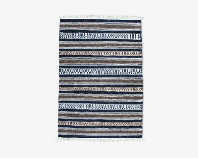 Rektangulært vævet tekstil med vandrette striber i blå, brune og hvide nuancer samt indviklede geometriske mønstre. Kanterne har hvide frynser på begge korte sider, hvilket giver et dekorativt udtryk.