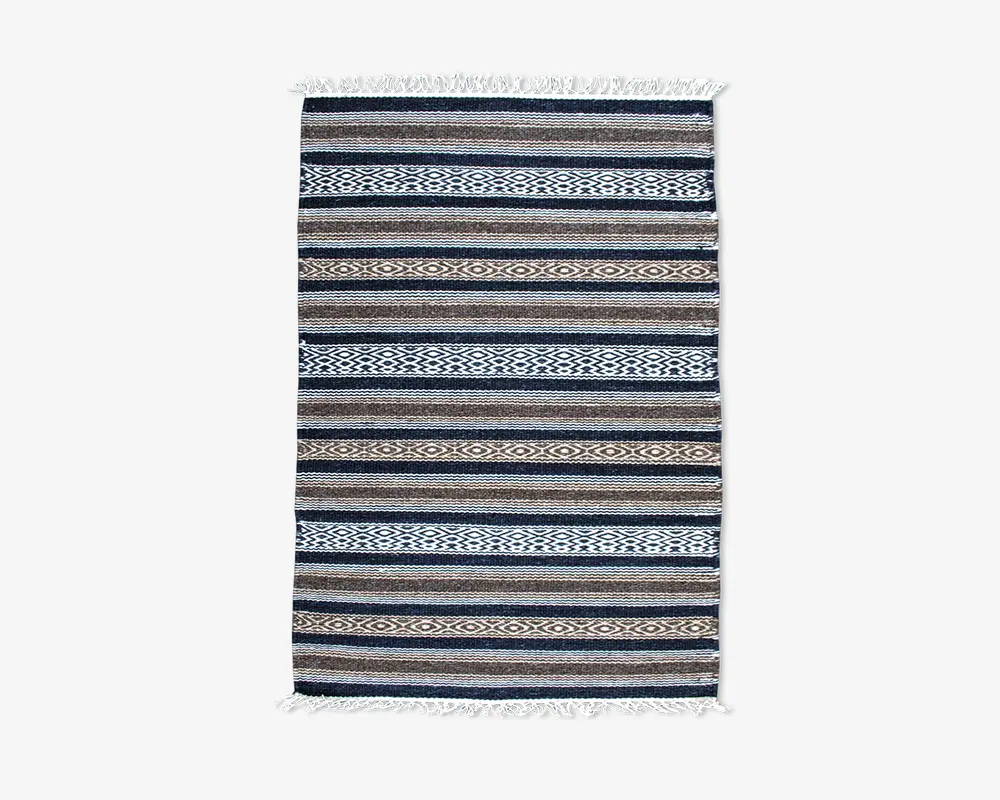 Rektangulært vævet tekstil med vandrette striber i blå, brune og hvide nuancer samt indviklede geometriske mønstre. Kanterne har hvide frynser på begge korte sider, hvilket giver et dekorativt udtryk.