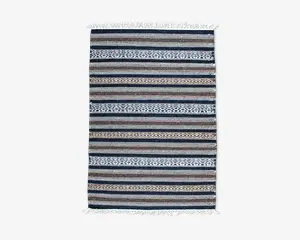 Rektangulært vævet tekstil med vandrette striber i blå, brune og hvide nuancer samt indviklede geometriske mønstre. Kanterne har hvide frynser på begge korte sider, hvilket giver et dekorativt udtryk.