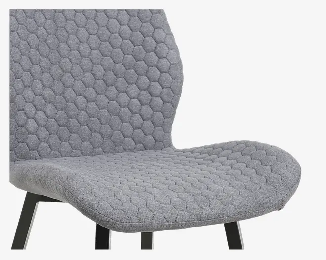 Nærbillede af en moderne stol med lysegråt stofbetræk og sekskantet quiltet mønster på både sæde og ryg. Stolen har et bredt, buet sæde og ryg samt skråtstillede sorte metalben.