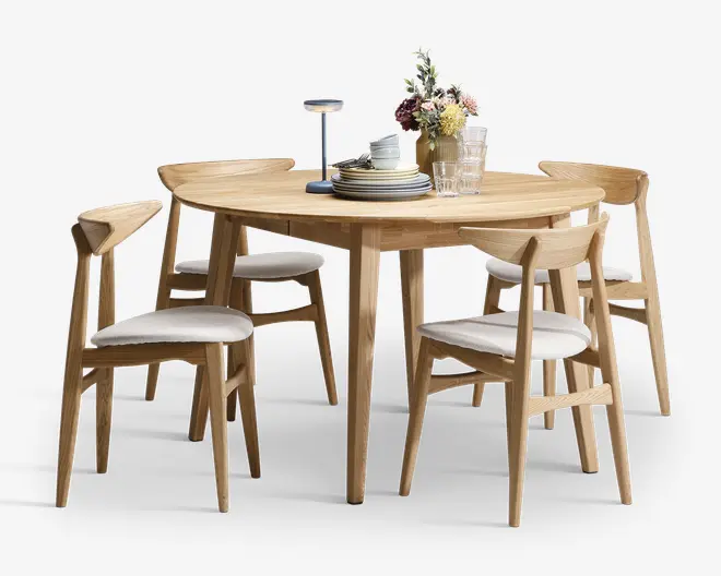 Et rundt spisebord lavet af lyst træ med fire matchende stole. Stolene har skrå ryglæn og hvide polstrede sæder for ekstra komfort. Bordet har koniske ben og er dekoreret med en stak tallerkener, glas og en vase med blomster, hvilket giver et moderne og indbydende udtryk.