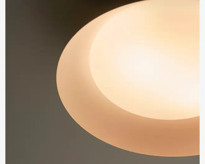 Nærbillede af en rund loftlampe med en varm hvid, lysende midte og en blød beige yderrand, som giver et glat, lagdelt udtryk mod en mørk baggrund.