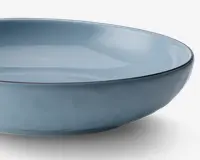 En lav, rund skål med en glat, blank overflade i en afdæmpet lyseblå farve. Skålens kant er let forhøjet og har en tynd, mørkere kant, der giver en diskret kontrast. Skålen har et minimalistisk design med rene linjer og ensartet farve.