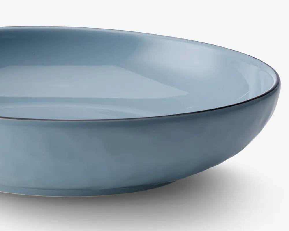 En lav, rund skål med en glat, blank overflade i en afdæmpet lyseblå farve. Skålens kant er let forhøjet og har en tynd, mørkere kant, der giver en diskret kontrast. Skålen har et minimalistisk design med rene linjer og ensartet farve.