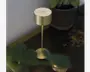 En moderne, minimalistisk lampe med en cylindrisk skærm og fod i guldfarvet metal, forbundet af en slank guldfarvet stang. Lampen har en touch-knap på toppen til betjening og et elegant, metallisk finish. Den står på en mørk træoverflade ved siden af en potteplante.