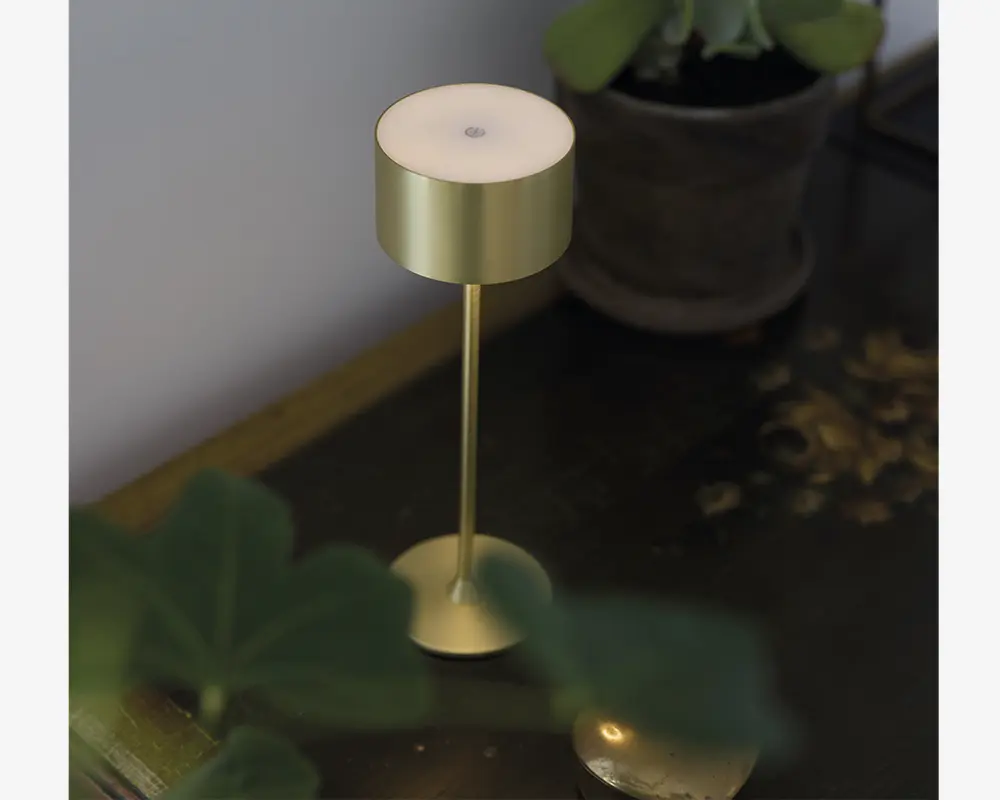 En moderne, minimalistisk lampe med en cylindrisk skærm og fod i guldfarvet metal, forbundet af en slank guldfarvet stang. Lampen har en touch-knap på toppen til betjening og et elegant, metallisk finish. Den står på en mørk træoverflade ved siden af en potteplante.