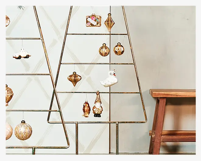Metallisk ramme formet som et juletræ med vandrette stænger, dekoreret med forskellige hængende ornamenter, herunder geometriske former, en svane, en bil og en julemandsfigur. Ornamenterne har guld-, hvid- og brunlige nuancer, og rammen står ved siden af en træbænk op ad en lys væg.
