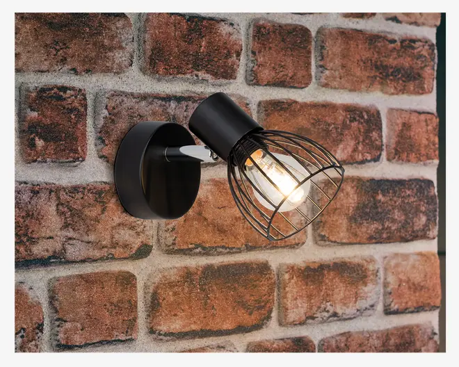 En vægmonteret lampe med mat sort finish og rund base. Lampen har en synlig pære omgivet af et sort trådbur, hvilket giver et industrielt udtryk. Lampen er monteret på en murstensvæg, og pæren udsender et varmt, indbydende lys.