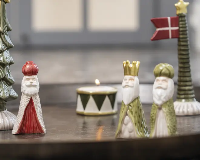 Små dekorative figurer af de tre vise mænd med hvide skæg og kåber i guld, rød og grøn, vist sammen med en stribet fyrfadsstage og et højt træ med en stjerne og et rødt flag.