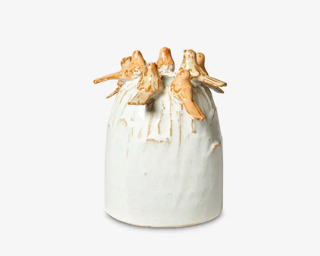 En keramikvase med mat, offwhite overflade, dekoreret med en gruppe skulpturerede fugle, der sidder rundt om kanten. Fuglene er glaseret i en varm, lysebrun farve, hvilket giver et dekorativt og tekstureret udtryk øverst på vasen. Vasen har en glat overflade med diskrete lodrette linjer og en let afrundet form.