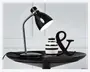 En elegant, moderne bordlampe med blank sort overflade og en fleksibel, kromfarvet svanehals, der gør det muligt at justere lysets retning. Lampen har en rund, solid fod og en konisk lampeskærm. Den står på et sort fad sammen med en lille, rund vase med sorte og hvide vandrette striber samt et dekorativt sort ampersand-symbol.