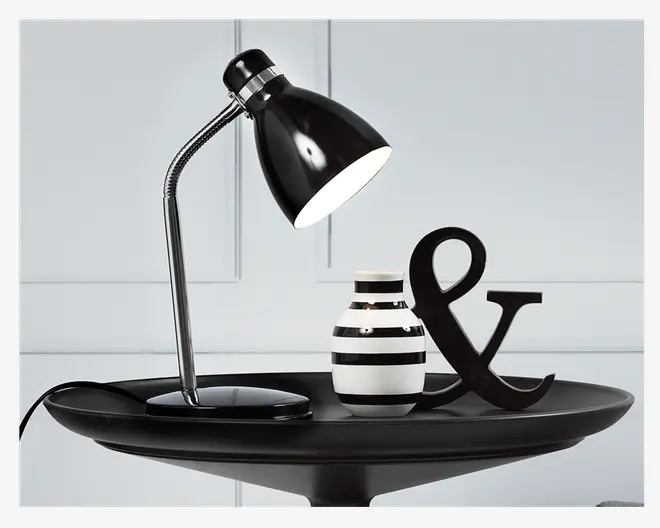 En elegant, moderne bordlampe med blank sort overflade og en fleksibel, kromfarvet svanehals, der gør det muligt at justere lysets retning. Lampen har en rund, solid fod og en konisk lampeskærm. Den står på et sort fad sammen med en lille, rund vase med sorte og hvide vandrette striber samt et dekorativt sort ampersand-symbol.