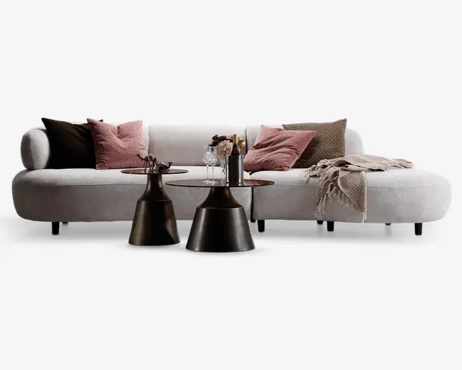 Moderne hjørnesofa med blødt, lysefarvet stofbetræk. Sofaen har afrundede kanter og en chaiselong til højre. Den er pyntet med flere pyntepuder i rosa, brun og beige nuancer samt et beige, tekstureret tæppe lagt over chaiselongen. Foran sofaen står to runde, mørke metalborde, begge med dekorative genstande som glas, en flaske og en lille vase med blomster.