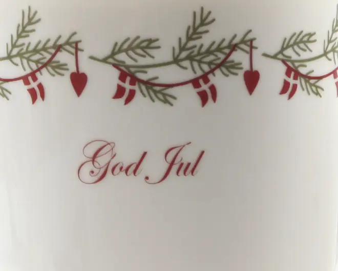 Billedet viser en hvid overflade med et festligt design øverst, bestående af grønne grangrene, røde guirlander, små røde flag og hængende røde hjerteornamenter. Under dekorationen står teksten 'God Jul' skrevet med elegant rød skrift, hvilket giver en julehilsen. Det samlede udtryk er klassisk og velegnet til juletema.