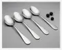 Fire identiske spiseskeer i rustfrit stål med blank, spejlende overflade ligger på række på en glat, lysegrå baggrund. Hver ske har et let buet skaft, der smalner ind og ender afrundet. Skeerne har et klassisk, enkelt design uden dekorationer. Tre blåbær ligger øverst til højre som størrelsesreference.