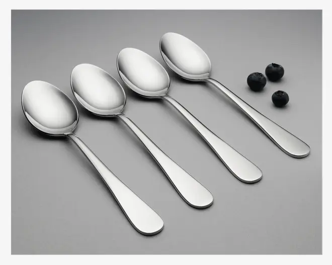 Fire identiske spiseskeer i rustfrit stål med blank, spejlende overflade ligger på række på en glat, lysegrå baggrund. Hver ske har et let buet skaft, der smalner ind og ender afrundet. Skeerne har et klassisk, enkelt design uden dekorationer. Tre blåbær ligger øverst til højre som størrelsesreference.