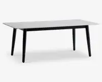 Rektangulært bord med en glat, hvid bordplade og fire let skråtstillede sorte ben. Designet er minimalistisk og moderne med rene linjer og en slank profil. Bordet virker solidt og rummeligt, velegnet til spisning eller arbejde.