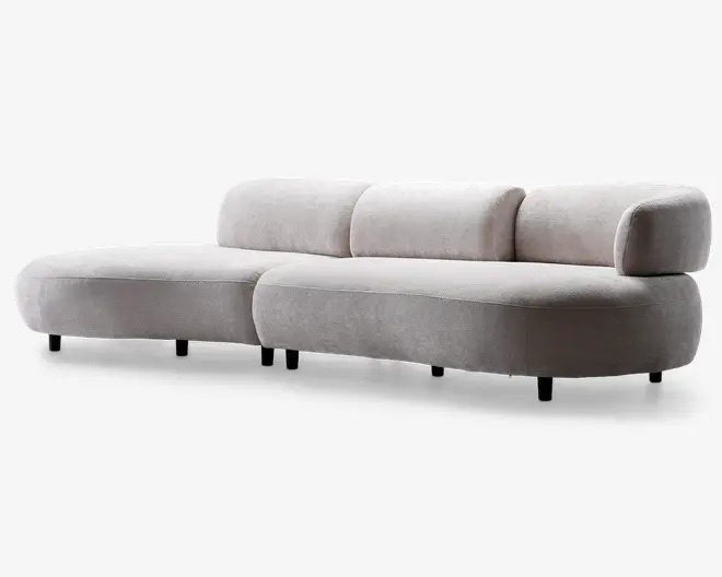Moderne sofa med lyst beige stofbetræk og et unikt buet design. Ryglæn og sædehynder er tykke og bløde, hvilket giver god komfort. Armlænet er kun på den ene side, hvilket giver sofaen et asymmetrisk og moderne udtryk. Sofaen står på korte, næsten usynlige ben, hvilket understreger det elegante og minimalistiske look.