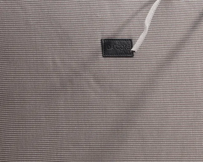 Nærbillede af et fint tekstureret, lysegråt stof med diskrete horisontale striber. Der er et lille, rektangulært sort mærke med mærkenavnet 'D4CORE' syet nær øverste højre hjørne, og en hvid snor er delvist synlig over mærket. Materialet fremstår glat og slidstærkt, velegnet til afslappet eller sportstøj.