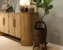 Rustikt træskab med slidt finish, to låger og lange lodrette greb, stylet med en bordlampe, dekorative vaser, en lysestage og en skulpturel figur ovenpå. En høj plante med store blade står i en mørk tøndeformet plantekasse på en lille træskammel ved siden af.
