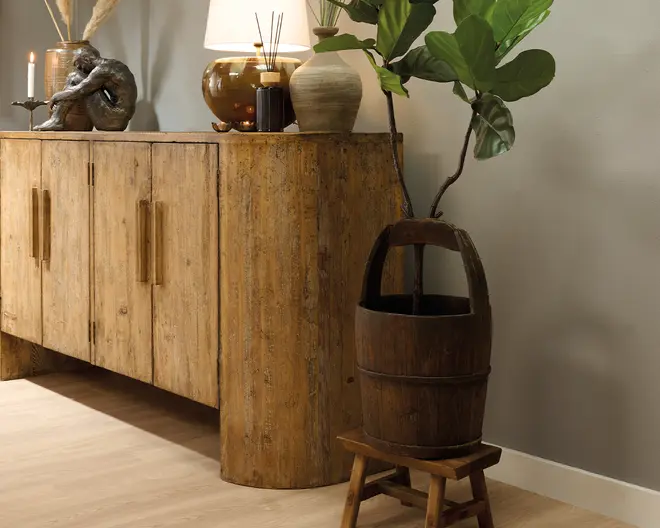 Rustikt træskab med slidt finish, to låger og lange lodrette greb, stylet med en bordlampe, dekorative vaser, en lysestage og en skulpturel figur ovenpå. En høj plante med store blade står i en mørk tøndeformet plantekasse på en lille træskammel ved siden af.