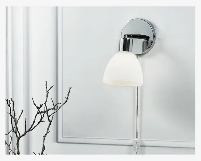 Væghængt lampe med rund, forkromet base og en nedadrettet, mat hvid glasskærm. Lampen har en synlig ledning, der hænger lige ned, og et moderne, minimalistisk design. Den forkromede overflade giver et elegant og reflekterende udtryk.