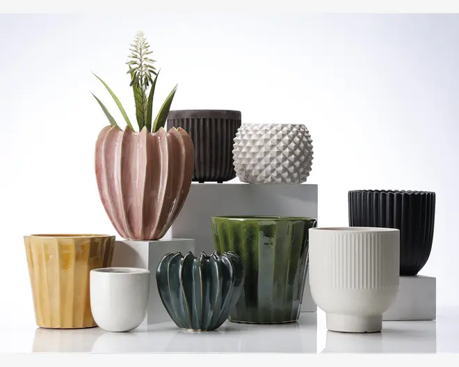 En samling keramiske urtepotter i forskellige former, størrelser og farver. Urtepotterne har forskellige overflader, herunder riflede, rillede, facetterede og glatte finish. Farverne spænder fra pastelrosa, sennepsgul og dybgrøn til blank hvid, mørkeblå og mat sort. Nogle potter har et afrundet, bølget design, mens andre er cylindriske eller har geometriske mønstre. Én potte indeholder en grøn plante med lange blade og en hvid blomst.