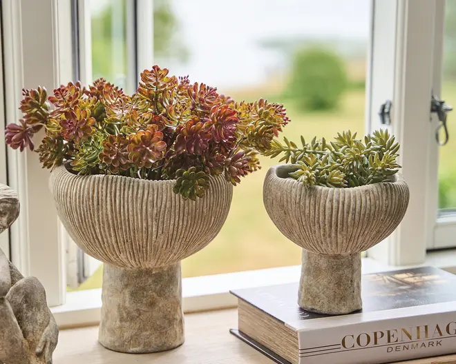 To teksturerede skåle i stenlook med ribbede, rillet sider, én stor og én mindre, begge fyldt med sukkulentlignende grønne planter. De står i en vindueskarm ved siden af en stak bøger med et blødt udendørs baggrundsview.