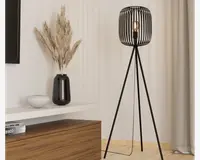 Høj gulvlampe med moderne design, sort metalskærm i bur-stil og tre slanke ben i tripod-form. Lampeskærmen er åben og viser pæren, hvilket giver et industrielt udtryk. Lampen står på et lyst trægulv ved siden af en hvid væg og gardin.
