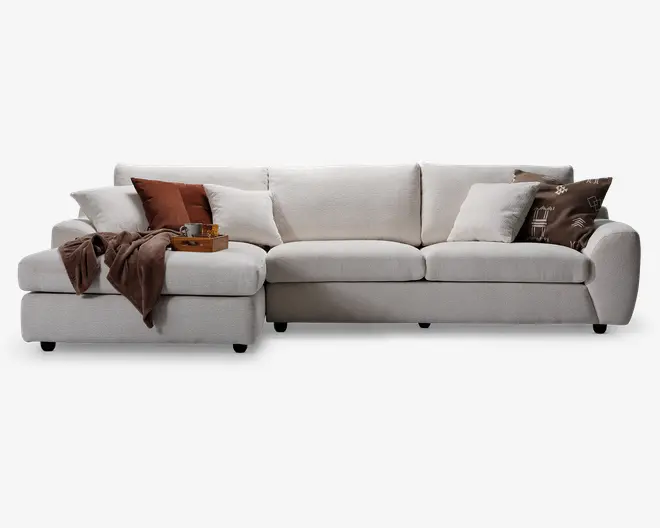 Stor L-formet sofa betrukket med lysebeige stof, med brede armlæn og dybe sædehynder. Sofaen er pyntet med flere pyntepuder i hvid, brun og mønstrede designs samt et brunt tæppe lagt over den ene side. En træbakke med en kop og småting står på chaiselongen, hvilket fremhæver det rummelige afslapningsområde.