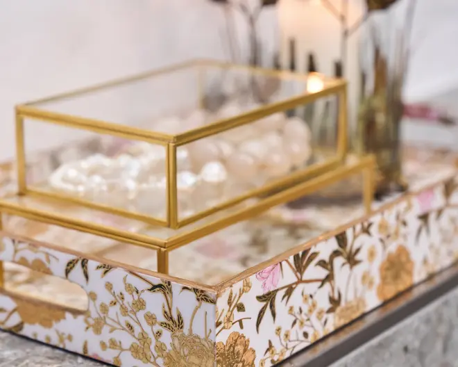 Rektangulær dekorativ bakke med udskårne håndtag på siderne, dekoreret med et hvidt baggrundsmønster af indviklede guld- og lyserøde blomster. Ovenpå bakken står en glaskasse med guldfarvede metalrammer, som indeholder en samling små, runde, gennemsigtige genstande. Det samlede udtryk er elegant og luksuriøst, velegnet til at organisere eller udstille ting på en stilfuld måde.