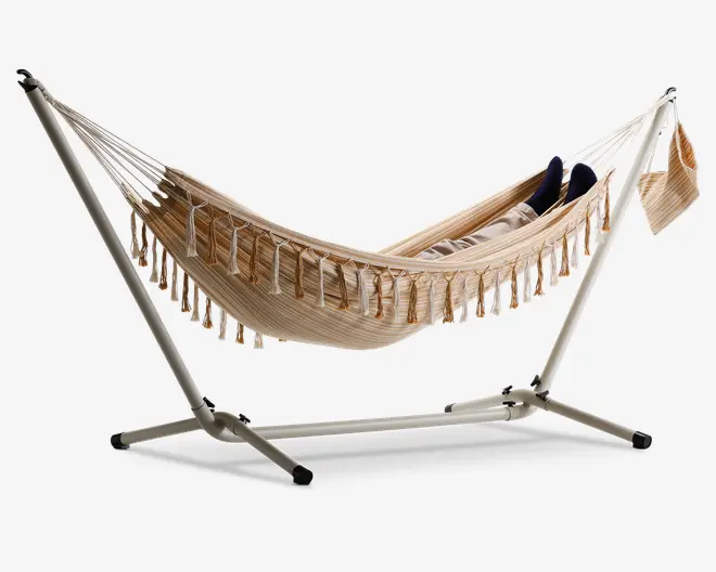 Beige og cremefarvet stribet hængekøje med dekorativ frynsekant langs siderne, ophængt på et robust metalstativ. Hængekøjen har et vævet stofdesign og en matchende stofpose fastgjort i den ene ende. Stativet har en pulverlakeret overflade og brede, stabile ben for god støtte.