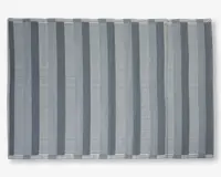 Rektangulært tekstil med skiftende lodrette striber i grå og hvide nuancer. Striberne varierer i bredde og mønster, hvor nogle er ensfarvede og andre består af tynde vandrette linjer, hvilket giver et visuelt tekstureret udtryk. Kanterne er pænt afsluttet med en enkel syet kant.