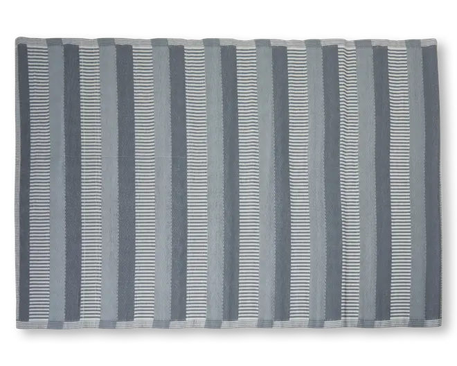 Rektangulært tekstil med skiftende lodrette striber i grå og hvide nuancer. Striberne varierer i bredde og mønster, hvor nogle er ensfarvede og andre består af tynde vandrette linjer, hvilket giver et visuelt tekstureret udtryk. Kanterne er pænt afsluttet med en enkel syet kant.