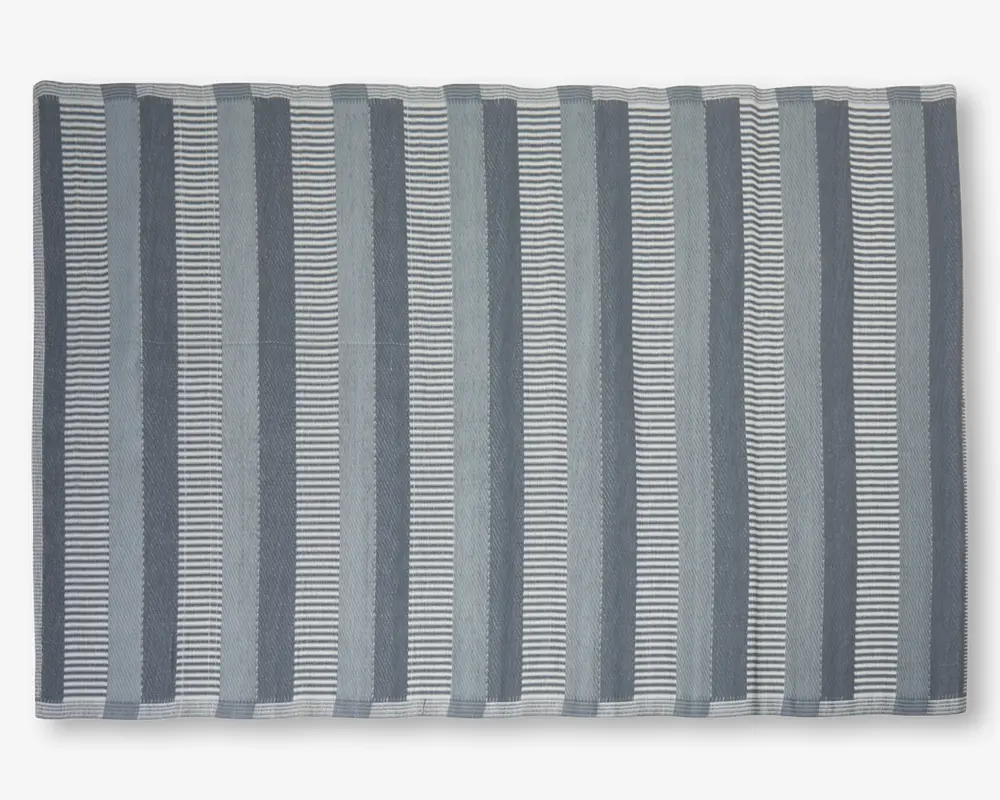Rektangulært tekstil med skiftende lodrette striber i grå og hvide nuancer. Striberne varierer i bredde og mønster, hvor nogle er ensfarvede og andre består af tynde vandrette linjer, hvilket giver et visuelt tekstureret udtryk. Kanterne er pænt afsluttet med en enkel syet kant.