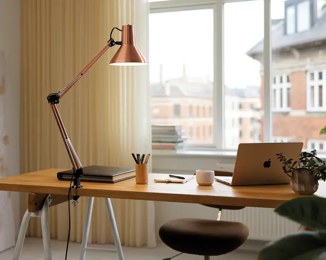 Et moderne arbejdsområde med et skrivebord i træ og en elegant, justerbar bordlampe i kobber. På skrivebordet står en lukket bærbar computer, en kop med flere kuglepenne, nogle løstliggende penne og blyanter, et keramikkrus og en potteplante. Skrivebordet står foran et stort vindue med cremefarvede gardiner, som lader masser af naturligt lys komme ind i rummet. Udtrykket er ryddeligt, organiseret og indbydende, med en komfortabel brun polstret stol.