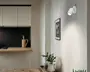 Minimalistisk hvid cylindrisk væglampe på rund bagplade, vinklet nedad og let udad, med et enkelt moderne udtryk.
