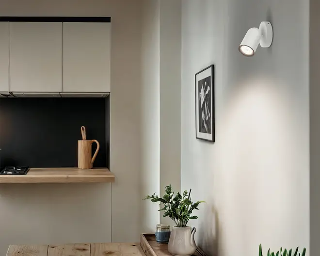 Minimalistisk hvid cylindrisk væglampe på rund bagplade, vinklet nedad og let udad, med et enkelt moderne udtryk.