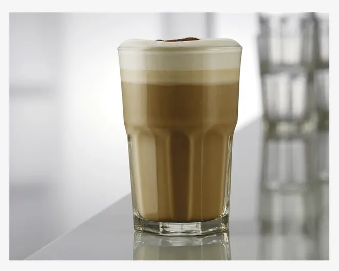 Et klart glas fyldt med en lagdelt kaffedrik, der har en dyb brun kaffebund og et tykt, cremet lag mælkeskum på toppen. Glasset har et facetteret design og står på en reflekterende overflade. Mælkeskummet er glat og let buet, med et strejf af kakaopulver drysset ovenpå.