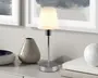 En moderne bordlampe med en slank, poleret kromfod og en lige, smal stang. Lampen har en enkel, konisk hvid lampeskærm, der spreder lyset blødt og giver en varm og indbydende glød. Designet er minimalistisk og moderne, hvilket gør lampen velegnet til forskellige indretninger. Lampen står på en lys overflade omgivet af dekorative genstande som en blank keramikvase og en rund, hvid kugle.