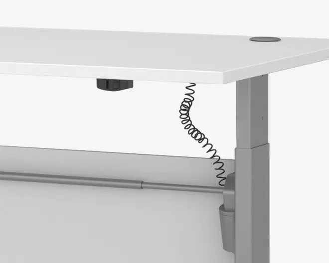 Nærbillede af et skrivebordshjørne med en hvid bordplade med afrundede kanter og kabelgennemføring. Bordet har et robust, rektangulært metalben i lysegrå finish, og under bordpladen ses en spiralformet strømkabel, der er tilsluttet en elektrisk stikkontakt monteret under overfladen. En vandret støttebjælke løber langs undersiden for ekstra stabilitet.
