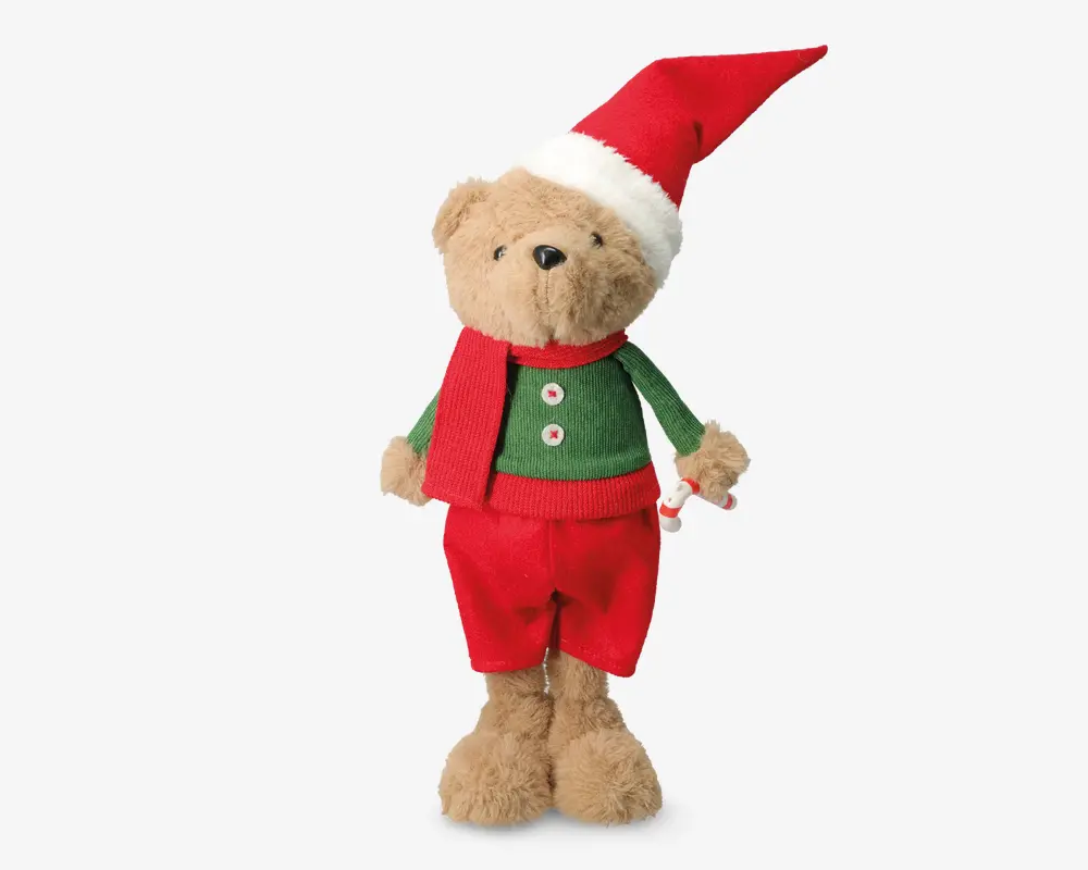 En bamse med lysebrun pels, iført et festligt outfit bestående af en rød nissehue med hvid kant, en grøn sweater med to hvide knapper, røde bukser og et matchende rødt tørklæde. Bamsen har et blødt og krammevenligt udseende og står oprejst.
