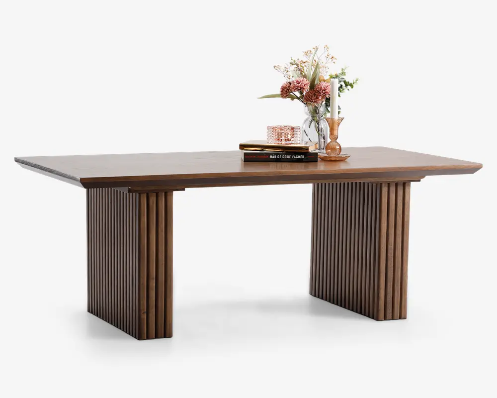 Rektangulært bord i træ med en glat, flad bordplade og to brede, lamelbeklædte ben. Træet har en varm, mellembrun finish. På bordet står der pyntegenstande, herunder en vase med blomster, to stablede bøger og en lysestage, hvilket giver et elegant og stilfuldt udtryk.