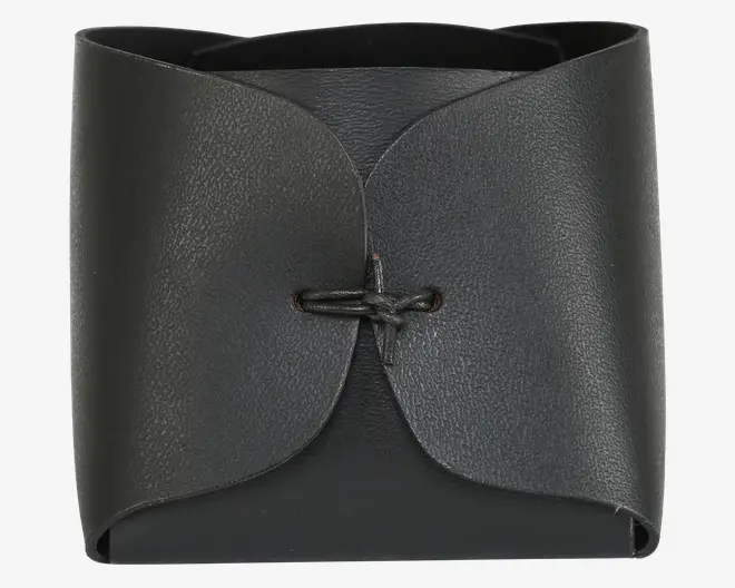 Et lille, kvadratisk produkt lavet af sort læder med en glat, mat overflade. Forsiden har to buede flapper, der overlapper hinanden og er fastgjort i midten med en enkel sort lædersnor, der er bundet gennem to huller. Kanterne er pænt afsluttede, og designet er minimalistisk og elegant.