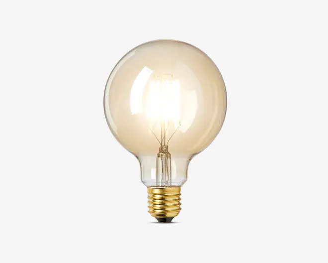 Klar, rund glaspære med synlig glødetråd indeni, der udsender et varmt hvidt lys. Pæren har en guldfarvet skruesokkel, som passer til almindelige fatninger. Designet er klassisk og dekorativt, hvilket gør den velegnet til lamper, hvor pæren er synlig.